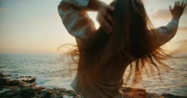 Okyanusun kenarında dans eden kadın dramatik günbatımı ışıklarıyla. Dansçı kameraya bakıyor.