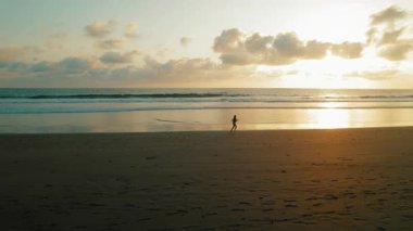 Gün batımında sakin okyanus dalgalarıyla kumlu sahilde koşan bir kadın..