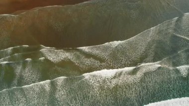 Okyanus dalgalarının üst görüntüsü koyu kumlu kıyıya doğru yuvarlanıyor desenli beyaz köpükle.