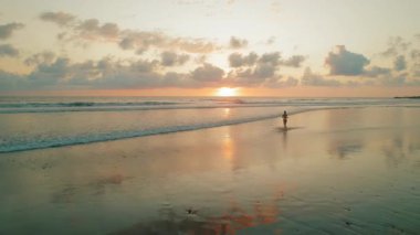 Gün batımında, ıslak sahilde yansımalarla okyanusa doğru yürüyen bir kadın..
