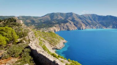 Kefalonya 'da dramatik uçurumlar ve mavi deniz manzaralı bir körfeze bakan Assos harabeleriyle kayalık yolda koşan bir kadın görülüyor..