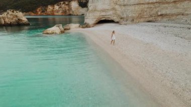 Turkuaz deniz suyu ve sahil kayalıkları olan sakin çakıl taşı plajlarında yürüyen bir kadın..
