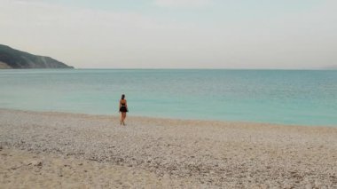 Sıcak bir yaz gününde, geniş çakıl taşı plajı boyunca sakin bir turkuaz denizin yanında yürüyen bir kadın..