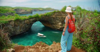 Sırt çantalı kadın yürüyüşçü Bali Endonezya 'daki Doğal Kemer Kırığı plajına karşı okyanus ve zevk teknesini izliyor.