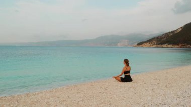 Woman meditating on calm turquoise beach in early morning light in Kefalonia Greece.