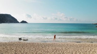 Güzel, uzun saçlı kadın turkuaz denizli beyaz çakıl taşı kumsalında duruyor..