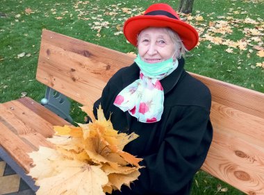 Koruyucu maskeli yaşlı bir kadın ve sonbahar yapraklı bir buket açık hava parkında eğleniyor..