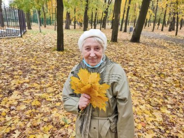 Koruyucu maskeli yaşlı bir kadın ve sonbahar yapraklı bir buket açık havada sonbahar parkında eğleniyor. COVID-2019 koronovirüs. Yaşlıların sağlığı. 80 yaşındaki bir kadının portresi.
