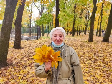 Koruyucu maskeli yaşlı bir kadın ve sonbahar yapraklı bir buket açık havada sonbahar parkında eğleniyor. COVID-2019 koronovirüs. Yaşlıların sağlığı. 80 yaşındaki bir kadının portresi.