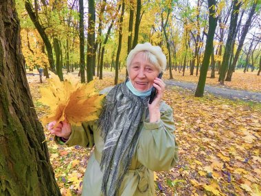 Koruyucu maskeli yaşlı bir kadın ve sonbahar yapraklı bir buket açık havada sonbahar parkında eğleniyor. COVID-2019 koronovirüs. Yaşlıların sağlığı. 80 yaşında cep telefonlu bir kadının portresi.