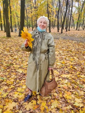 Koruyucu maskeli yaşlı bir kadın ve sonbahar yapraklı bir buket açık havada sonbahar parkında eğleniyor. COVID-2019 koronovirüs. Yaşlıların sağlığı. 80 yaşındaki bir kadının portresi.