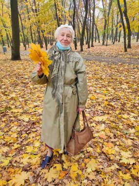 Koruyucu maskeli yaşlı bir kadın ve sonbahar yapraklı bir buket açık havada sonbahar parkında eğleniyor. COVID-2019 koronovirüs. Yaşlıların sağlığı. 80 yaşındaki bir kadının portresi.