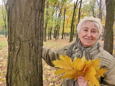 Gri saçlı, gülümseyen yaşlı kadın sonbahar parkında. Mutlu yaşlılık, doğa yürüyüşü, olumlu duygular..