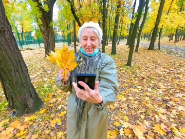 Koruyucu maskeli yaşlı bir kadın ve sonbahar yapraklı bir buket açık havada sonbahar parkında eğleniyor. COVID-2019 koronovirüs. Yaşlıların sağlığı. 80 yaşında cep telefonuyla selfie çeken bir kadının portresi.