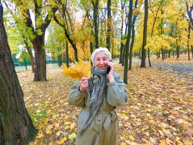 Koruyucu maskeli yaşlı bir kadın ve sonbahar yapraklı bir buket açık havada sonbahar parkında eğleniyor. COVID-2019 koronovirüs. Yaşlıların sağlığı. 80 yaşında cep telefonlu bir kadının portresi.