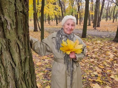 Gri saçlı, gülümseyen yaşlı kadın sonbahar parkında. Mutlu yaşlılık, doğa yürüyüşü, olumlu duygular..