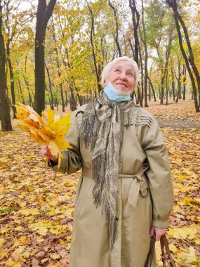 Koruyucu maskeli yaşlı bir kadın ve sonbahar yapraklı bir buket açık havada sonbahar parkında eğleniyor. COVID-2019 koronovirüs. Yaşlıların sağlığı. 80 yaşındaki bir kadının portresi.
