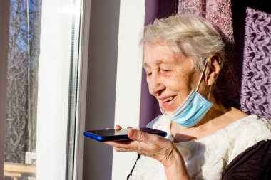 Akıllı telefonlu büyükannem cihazla sesli mesaj bırakıyor. Olgun kadın cep telefonunu sanal dijital ses asistanıyla kullanıyor. Covid-19, sağlık, salgın - virüsten korunmak için evde kıdemli