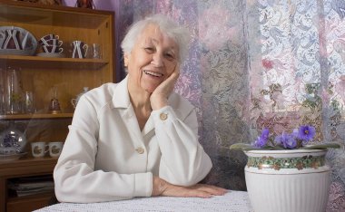 Yaşlı, yalnız, mutlu kadın evinin penceresinin yanında çiçeklerle oturuyor.