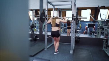 Formda bir kadın spor salonunda halterle ayna karşısında çömeliyor.