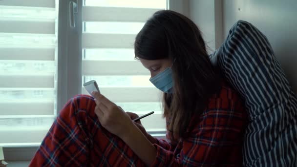 Девушка сидит у окна и рисует во время пандемии — стоковое видео
