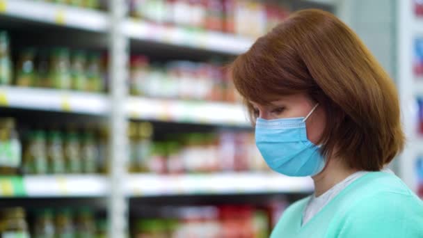 Mujer con máscara mirando estantes con comida enlatada en el supermercado — Vídeos de Stock