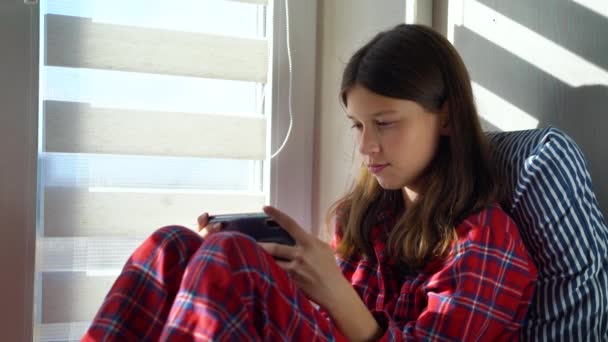 Девушка в пижаме сидит у окна и играет в онлайн игру — стоковое видео