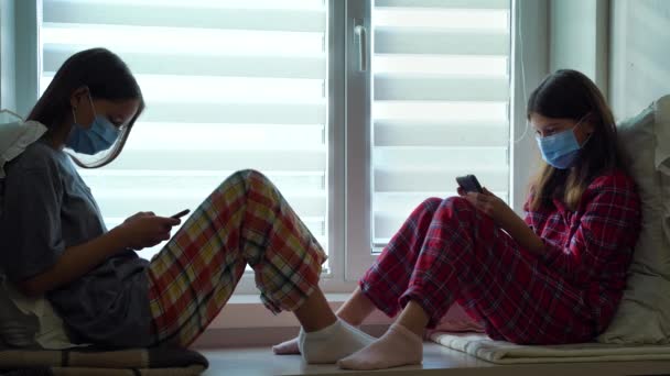 Sisters in masks sitting by window with smartphones — Stock Video