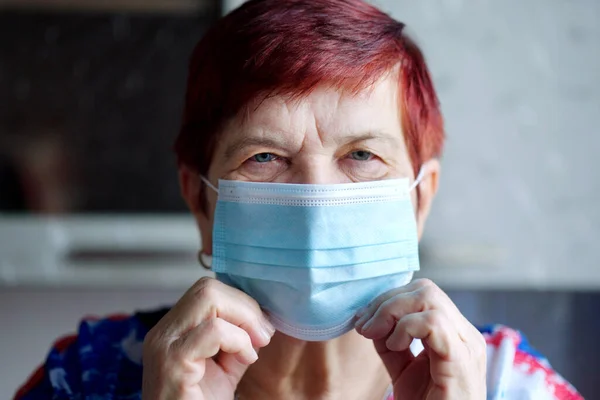 Senior woman putting on medical mask and looking at camera. Closeup aged person staying at home and social distancing during lockdown. Concept of healthcare