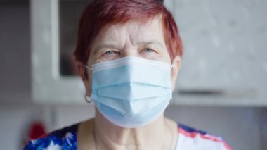Sağlık maskeli yaşlı kadın kameraya bakıyor.