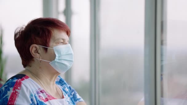 Ηλικιωμένη γυναίκα με μάσκα κοιτάζει έξω από το παράθυρο — Αρχείο Βίντεο
