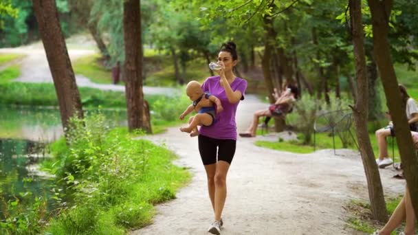 Mom carrying baby and drinking water after workout in park — Stockvideo