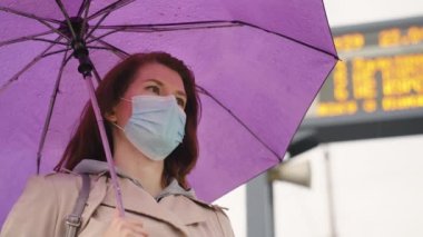 Otobüs durağında elinde şemsiyeyle duran maskeli kadın.