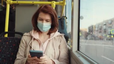 Otobüste akıllı telefon kullanan maskeli bir kadın.