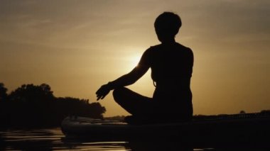 Gün batımında SUP tahtasında yoga yapan kadın silueti.