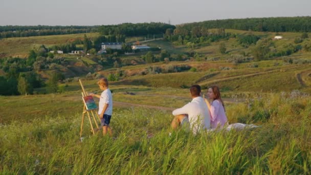 Сім'я відпочиває на пагорбі і малює хлопчика на мольберті — стокове відео