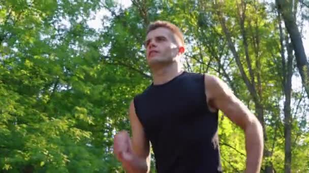 Slow motion young man jogging under green trees — ストック動画