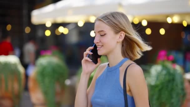 Blonde woman talking on phone while walking past street cafe — Stock Video