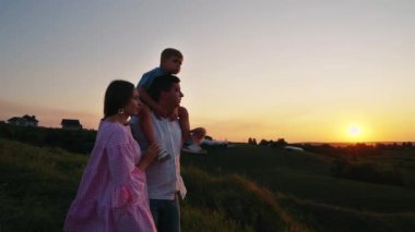 Ebeveynler gün batımında tepede durur, küçük çocuk babasının omuzlarında oturur, kır evleri arka planda kalır. Pan güzel manzarayı seyrederken ailesini vurdu. Bağlanma kavramı