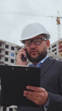 Kasketli ve takım elbiseli sakallı adam telefonda çok katlı binalara karşı konuşuyor. İnşaat alanında inşaat mühendisi.
