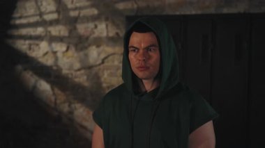 Kapüşonlu terli sporcu, ciddi bir ifadeyle doğrudan kameraya bakıyor. Huysuz spor salonunda, desenli duvarlar ve düşük ışıklandırmayla.