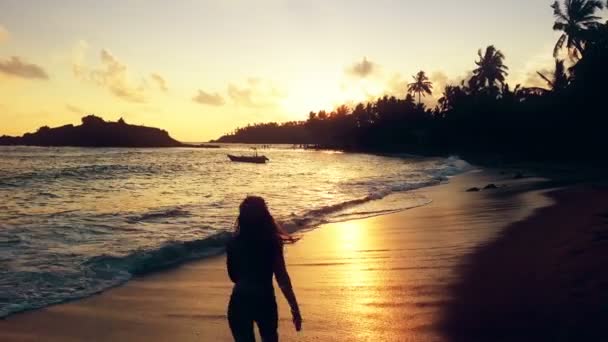 Lykkelig kvinne som nyter solnedgang på en tropisk strand, danser av glede i havbølger – stockvideo