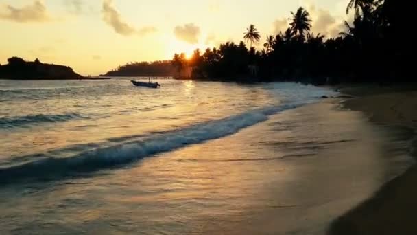 Store bølger skyller opp på sandstranden ved solnedgang, ensomme båter flyter i vann – stockvideo