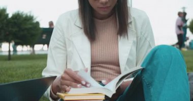 Kitap okuyan, parkta oturan, üniversite eğitimi alan, bilgili melez bir kadın.