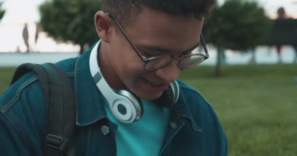 Hombre millennial en gafas con auriculares jugando juego, sentado al aire libre — Vídeos de Stock