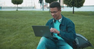 Gözlüklü siyah bir genç dışarıda dizüstü bilgisayarda yazı yazıyor, internette ödev yapıyor.