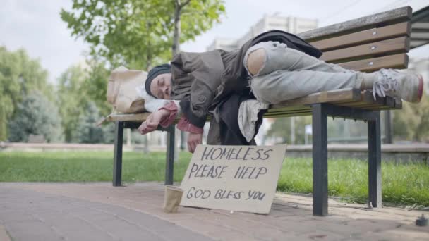 Женщина-беженка лежит на скамейке, бездомный картон поет на земле, жертва — стоковое видео