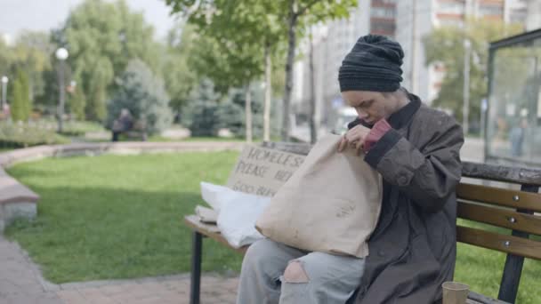 Бездомная женщина ищет еду в грязном мешке, сидит на скамейке, выживает — стоковое видео