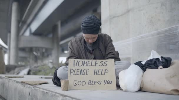 O femeie tristă stând pe pământ cu un semn fără adăpost, o persoană cu dizabilități — Videoclip de stoc
