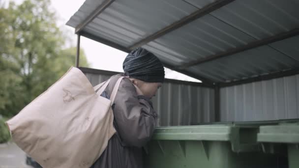 Bezdomna kobieta z brudną torbą, szukająca jedzenia w kontenerze na odpady. — Wideo stockowe